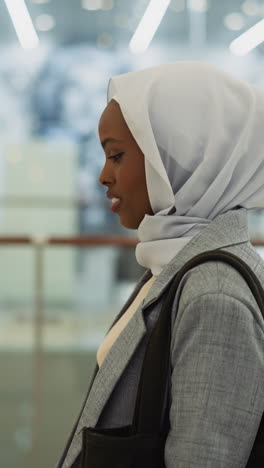 happy african-american lady with chiffon hijab walks along hall in shopping mall side view slow motion. muslim woman with handbag does shopping in center