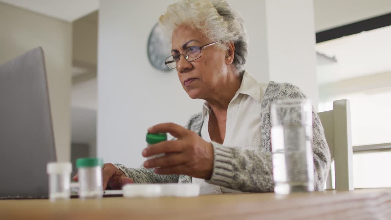 African american senior woman holding empty medication container while using laptop at home