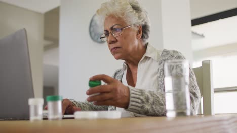 mujer mayor afroamericana sosteniendo un contenedor de medicamentos vacío mientras usa una computadora portátil en casa