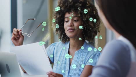 Animation-of-globe-of-icons-over-two-diverse-female-office-colleagues-discussing-over-a-document