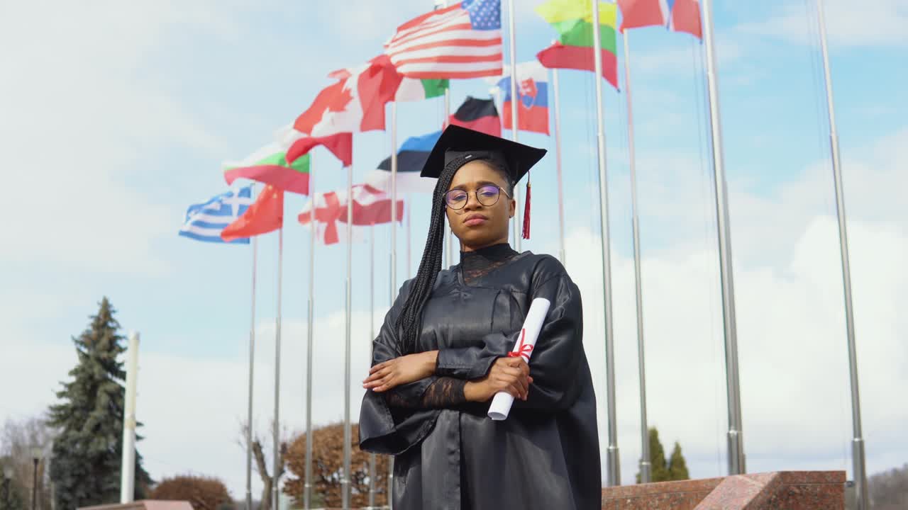 una joven afroamericana se alegra con un diploma de educación superior en la mano de pie contra el cielo y las banderas de diferentes países revoloteando en el viento.