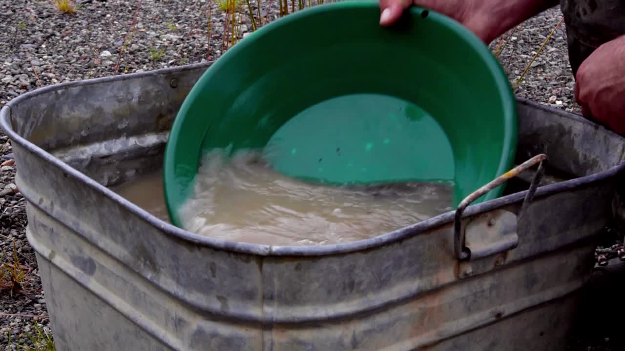 Gold panning black fine grunge, silt, sand, clay, and gravel alluvial deposits in green trap pan, wetting, soaking, agitating and washing in brown tin bucket of murky water, static close up portrait