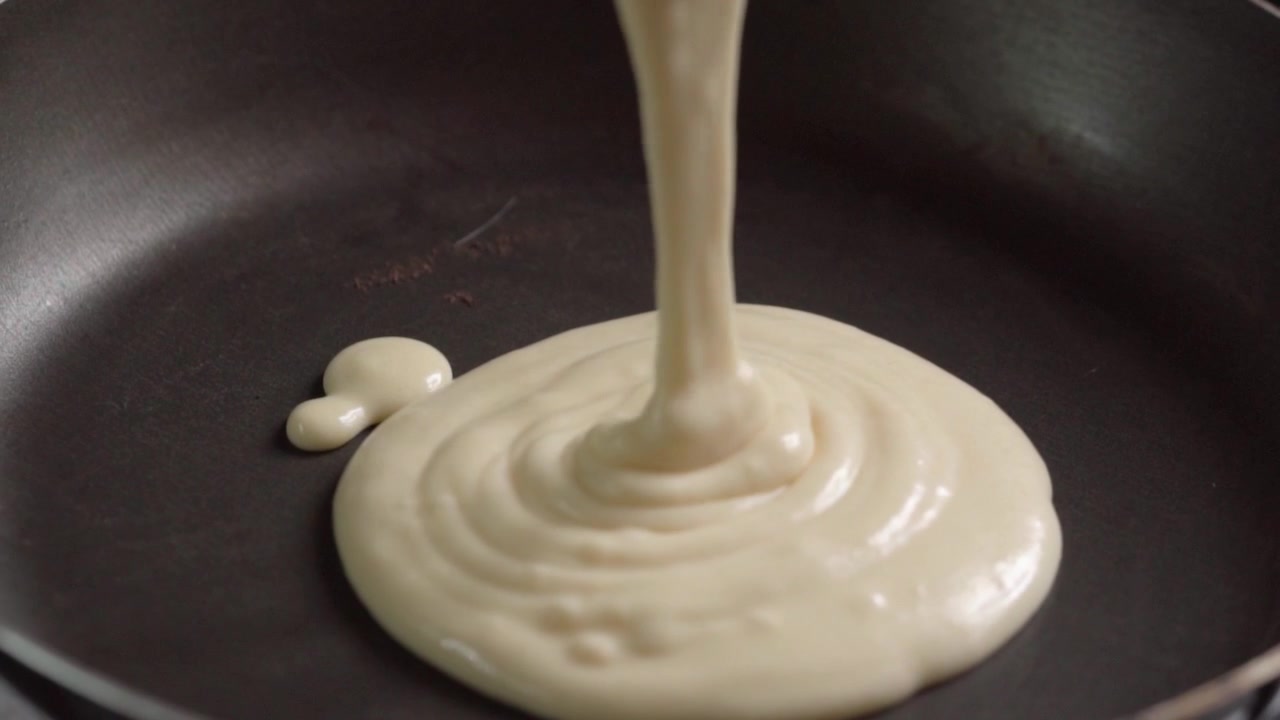 Close up of pancake batter pouring onto frying pan during breakfast preparation. Smooth batter spreading across heated surface showcasing homemade cooking and culinary process.