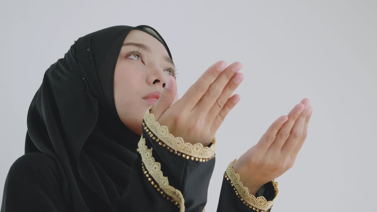 무슬림 아시아 여성은 검은 옷을 입고 히자브를 고 집 안의 거실에서 하나님께 축복을 구하기 위해 손을 들고 행복하게 미소 짓고 있습니다.