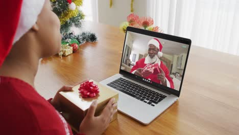 Afroamerikanische-Frau-Mit-Weihnachtsmütze-Nutzt-Laptop-Für-Einen-Weihnachtsvideoanruf-Mit-Dem-Weihnachtsmann-Auf-Dem-Bildschirm