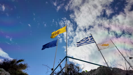 three flags fluttering against a vibrant sky with clouds: a blue flag, the greek flag, and a yellow flag with an emblem