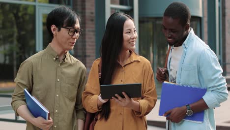 three asian and african american students standing near the university and looking at the tablet while discussing something