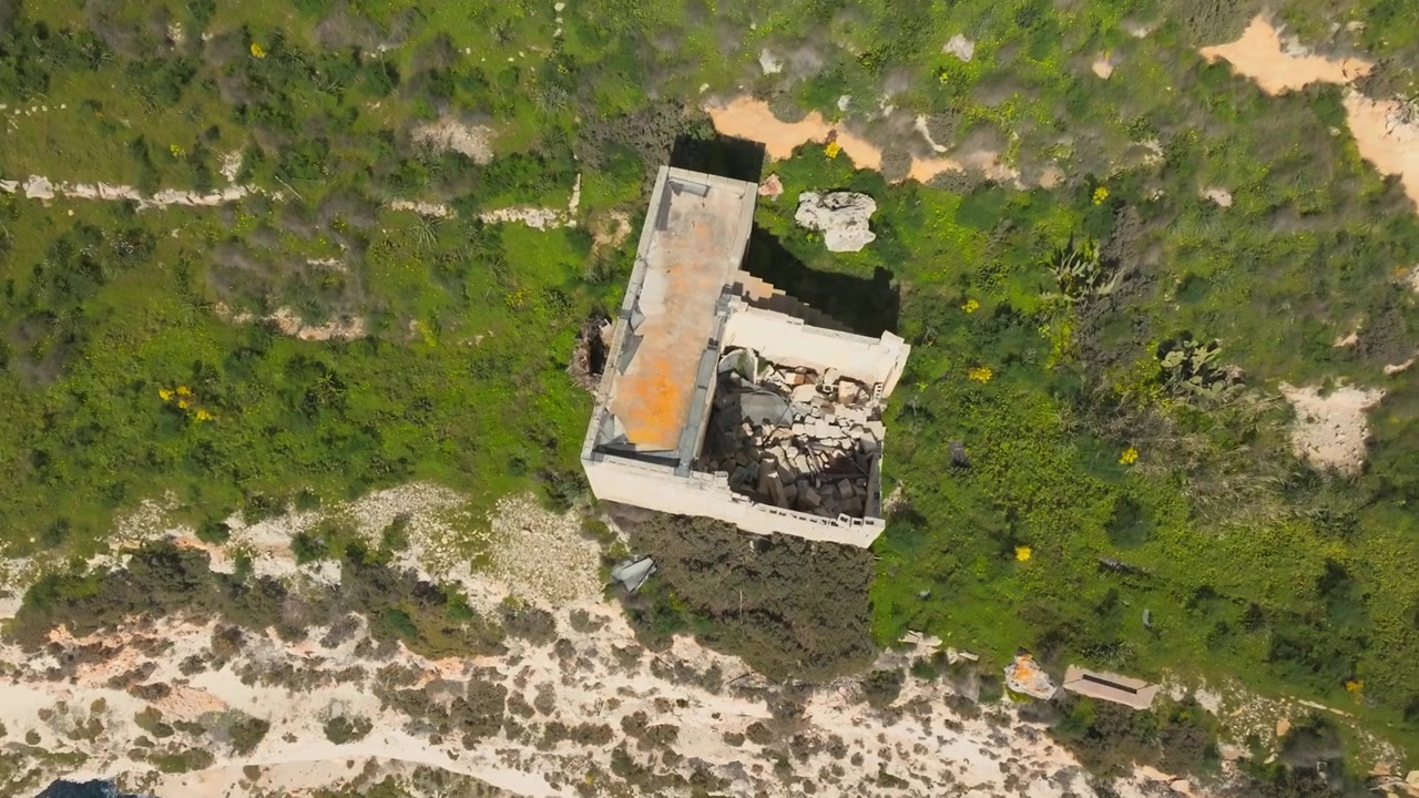 En naturskjønn klippe med grønn vegetasjon og en forlatt steinbygning har utsikt over et dypt blått hav under en delvis skyet himmel, med naturlig bevegelse av vann og skyer