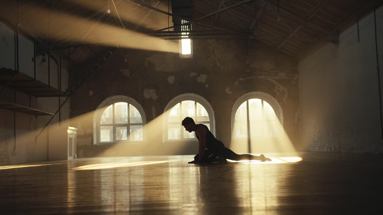 A man in a black sports summer uniform is doing stretching exercises in a gym that is lit by the rays of the sun