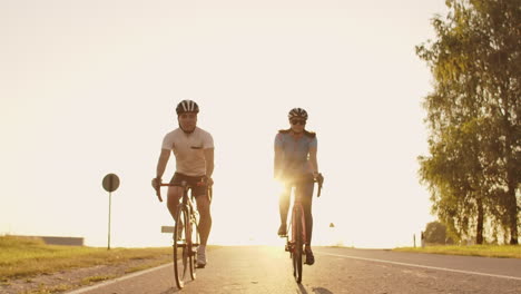 Un-Hombre-Y-Una-Mujer-Montan-Bicicletas-Deportivas-En-La-Carretera-Al-Atardecer-Con-Equipo-Y-Cascos-Protectores-En-Cámara-Lenta-A-120-Fps.