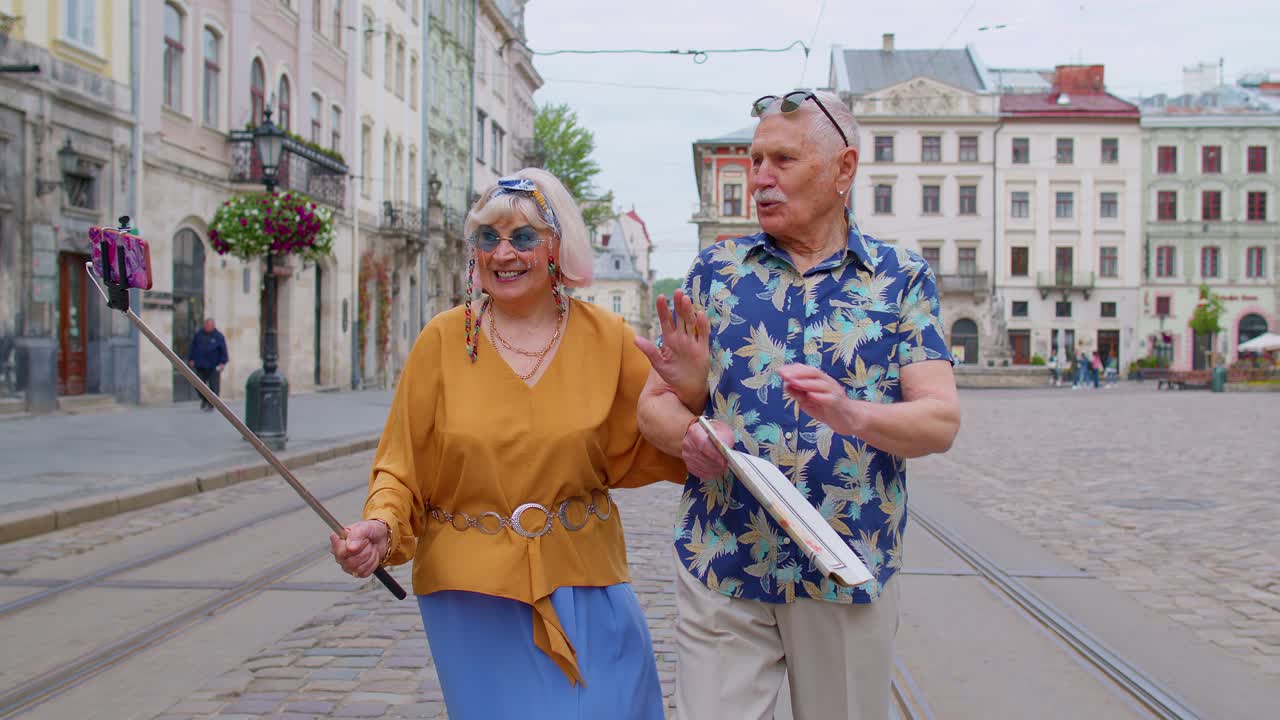 Senior old tourists man with woman walking in city with smartphone on selfie stick and taking photos