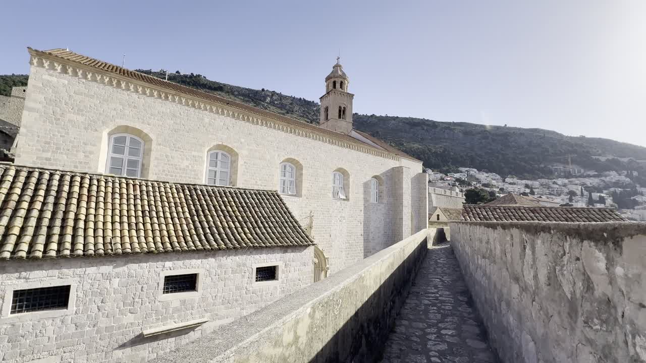 Lenta ripresa cinematografica a piedi lungo la sezione nord-orientale delle mura storiche della città di Dubrovnik durante le prime ore dell'alba. Luce calda che illumina l'architettura in pietra calcarea e storni che sorvolano la città