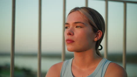 close-up of young lady with thoughtful expression as sunlight reflects off her face, sitting near iron railing during sunset, creating a moody atmosphere with blurred background