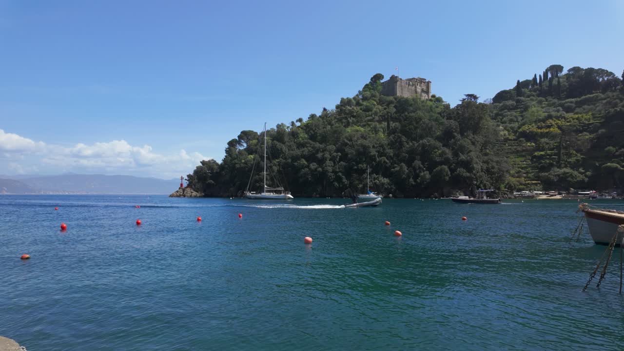 Inquadratura grandangolare di barche a vela che scivolano su un mare blu cristallino vicino a una collina verde lussureggiante con un castello in pietra sotto la luce del giorno intensa, riflessi vivaci sull'acqua e un ricco paesaggio costiero