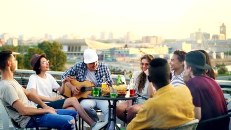 joyful musician is playing the guitar with his friends singing and laughing sitting on rooftop at table. entertainment, musical instruments and leisure concept.