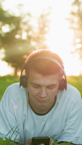 close up de artista de música acostado en la hierba con auriculares asintiendo con la cabeza a la música mientras usa el teléfono, la luz del sol mejora el brillo alrededor de la cabeza con un fondo borroso suave