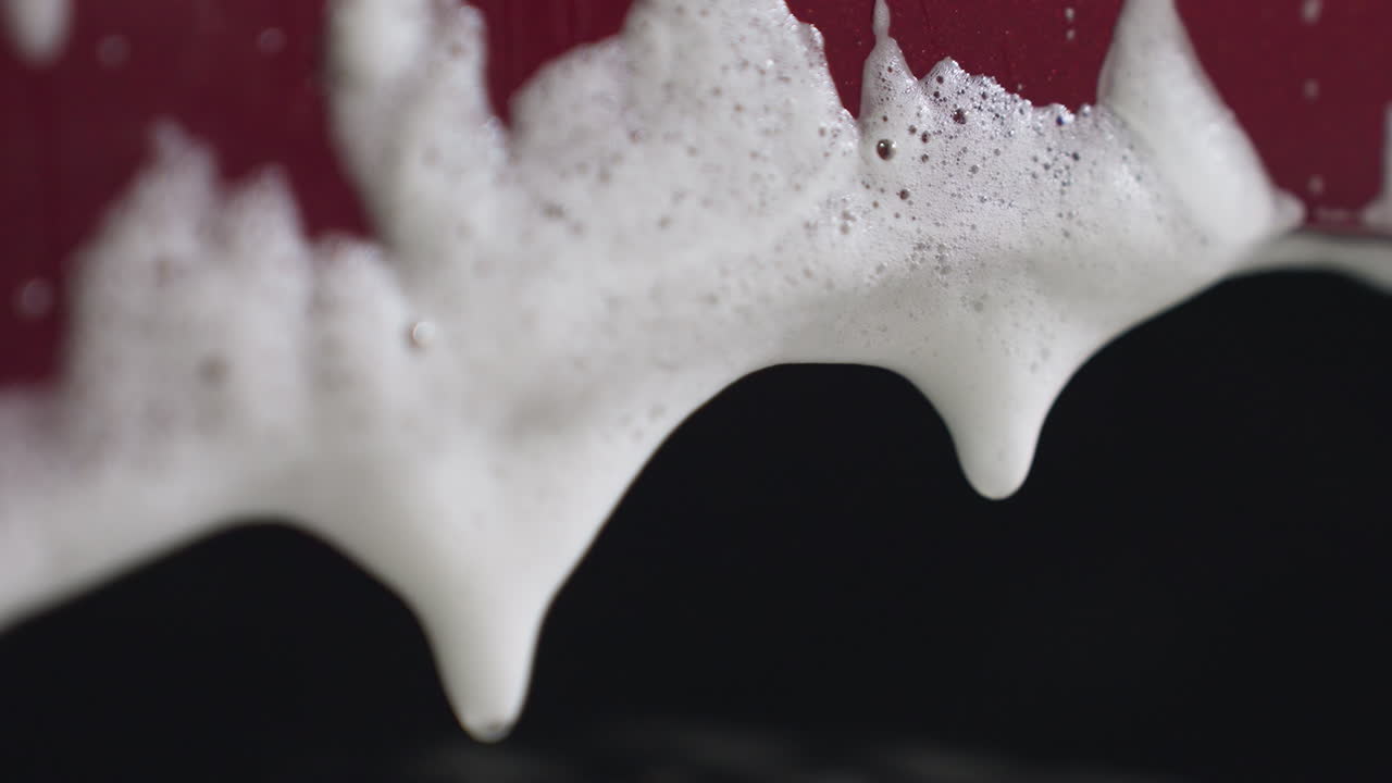 Close up of soapy foam forming drop and dripping from car body during washing process with water droplets creating dynamic texture and fluid movement against dark blurred background