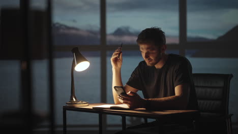 un hombre pensativo sentado en una mesa con una lámpara de escritorio y un teléfono móvil. el día oscuro es crepúsculo contra el fondo de la ventana un hombre triste está pensando en el trabajo mientras está sentado en casa.