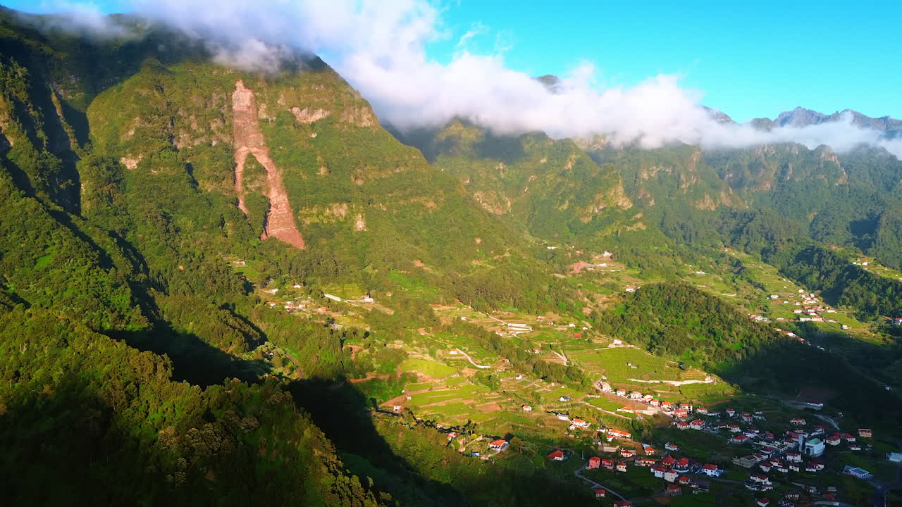 Little town situated in the green valley at the foot of high mountains. White cloudscape covering the tops of the rocks.