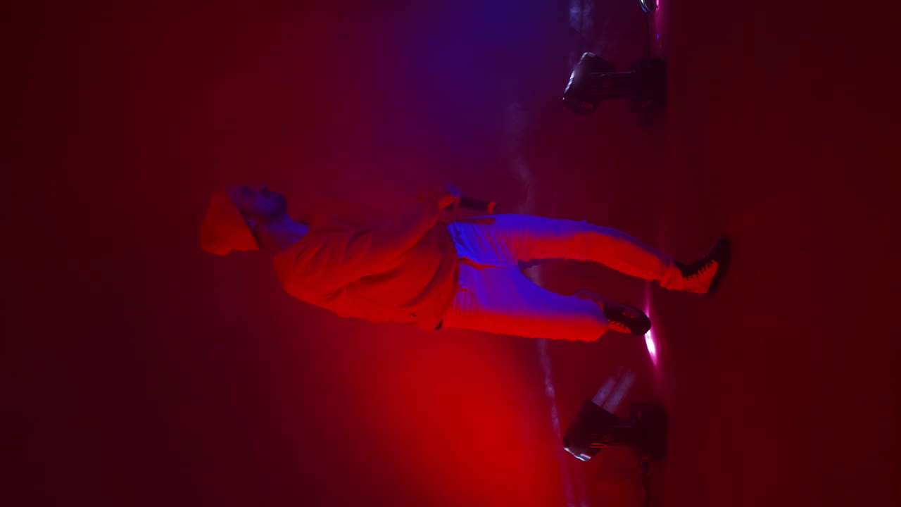 Vertical video. A rap artist sings into a microphone and dances in neon light and bright strobe lights in the smoke. The hip-hop singer sings energetically against changing background of the Studio