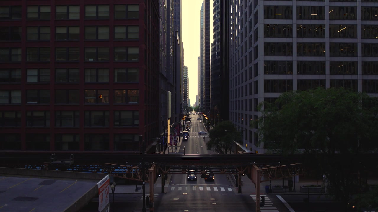 Vista drone su una strada del centro di Chicago sotto i binari sopraelevati della CTA, incorniciata da grattacieli, traffico, alberi e una calda luce serale