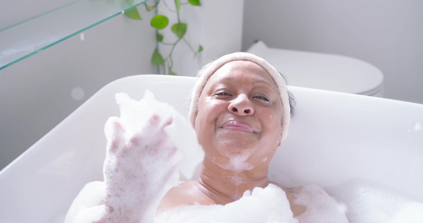 Senior African American woman reclining in bathtub lifting foam, blowing bubbles while relaxing