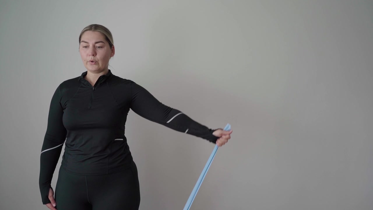 Volwassen vrouw in zwarte sportkleding voert een staande zijwaartse schouderheffing uit met een weerstandsband in een eenvoudige binnenruimte