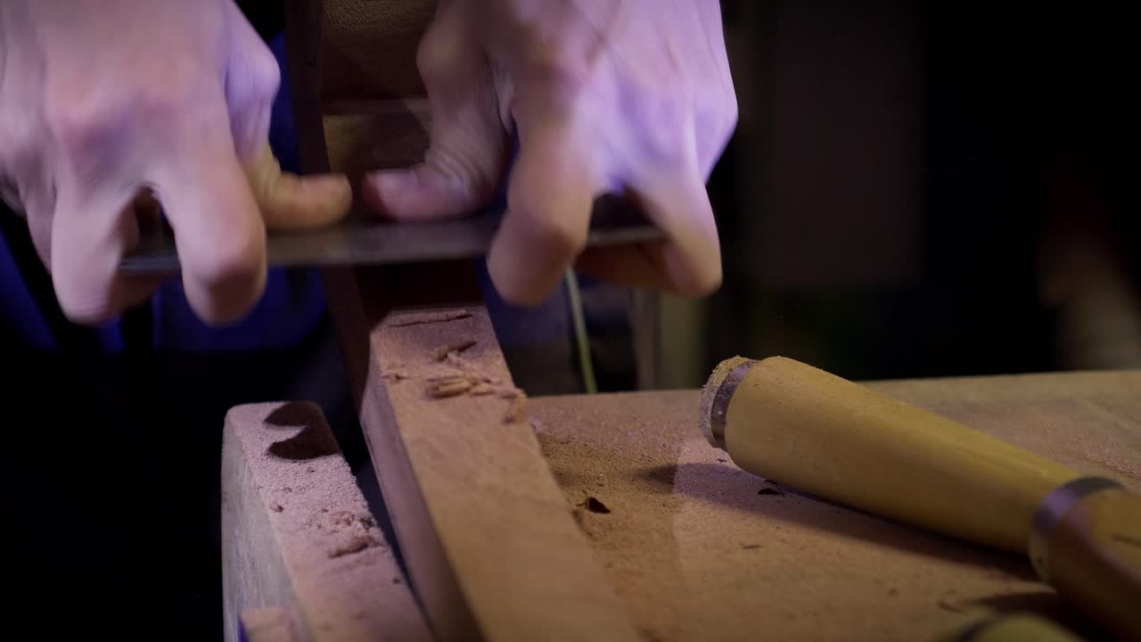 Woodworker using a metal scraper to refine the shape of a curved timber piece on a workbench. Slow motion close up
