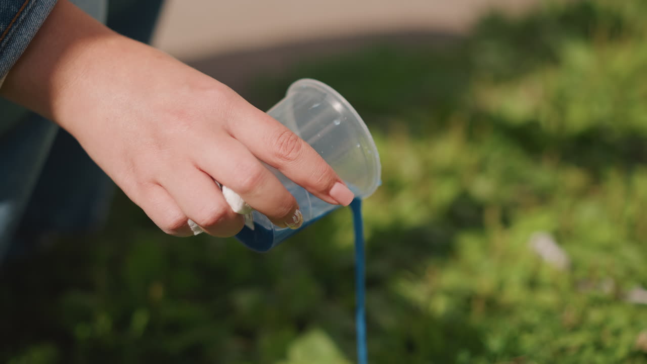 Blue paint spills onto lawn. Youthful hand pours colorful pigment into small blue container. Small child uses fingers to splash bright blue paint onto grassy surface during summer activity
