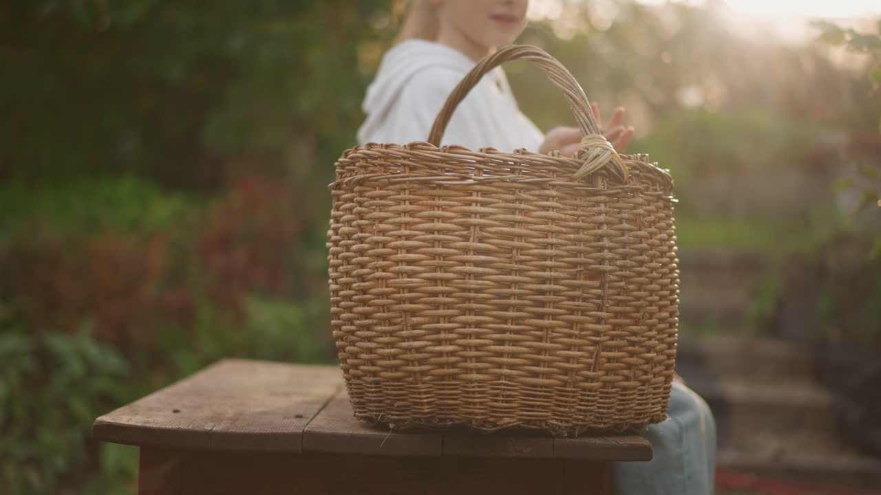 Cesto intrecciato illuminato dal sole su una panca, ragazza seduta che gira il manico di una grande borsa di paglia, luce dorata soffusa in controluce, gradini del giardino e vegetazione sfocata, atmosfera pastorale e tranquilla, tonalità autunnali stagionali.