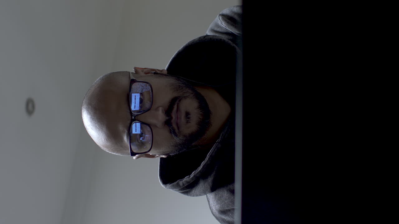 A middle-aged male wearing a hoodie is sitting in front of a laptop, browsing some videos while he has eyeglasses on