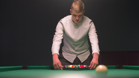 player in and gray trousers carefully arranges colorful billiard balls in triangle on green pool table. overhead lights cast warm glow as he focuses with calm concentration in stylish lounge