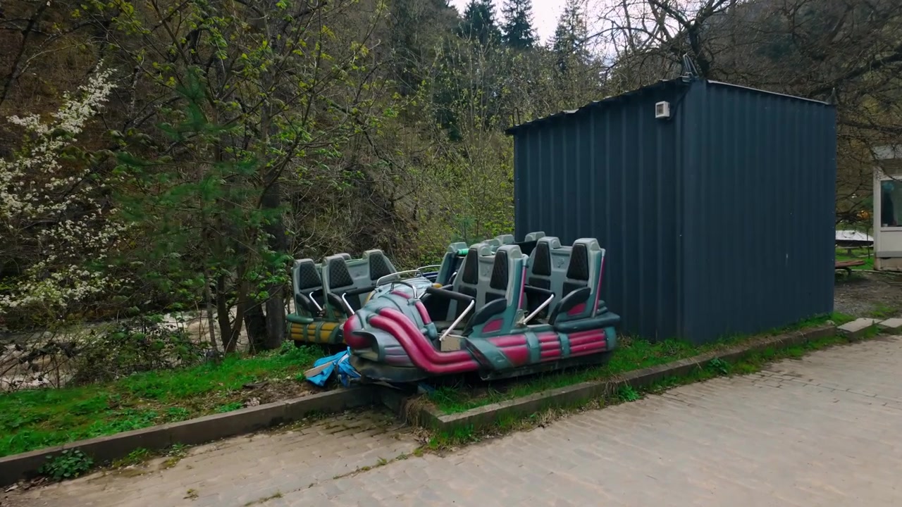 Paved path runs alongside a river, with discarded amusement park ride parts, a dark metal container, and a white booth. Surrounded by lush greenery and trees in an outdoor setting.
