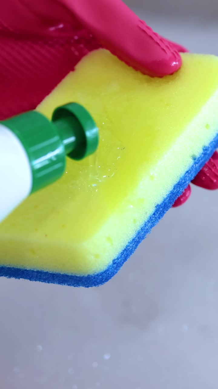 A hand in a red glove applies dish soap to a yellow sponge. Bright lighting enhances the cleaning process