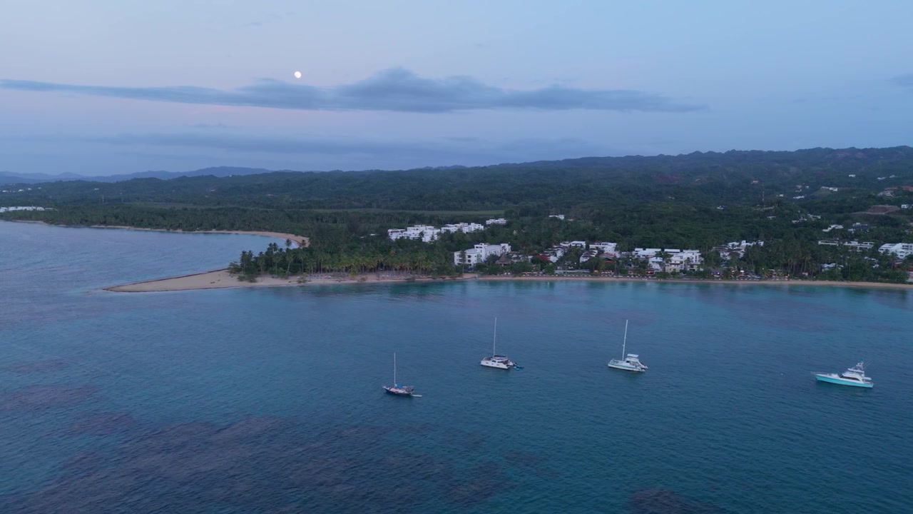 Ripresa aerea con drone che sorvola una baia calma con barche a vela e yacht ancorati, rivelando una punta sabbiosa tropicale e colline lussureggianti sotto un cielo crepuscolare illuminato dalla luna