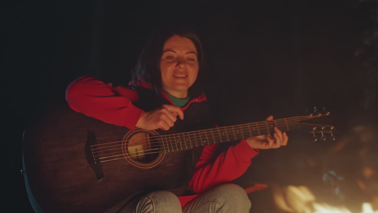 Panoramabillede af en camper, der spiller guitar ved et bål, med livlige sangudtryk, optimistisk guitarspil, iført afslappet jakke, siddende på en campingstol, med gnister og glød, en livlig udendørs optræden og en venlig lejrstemning.