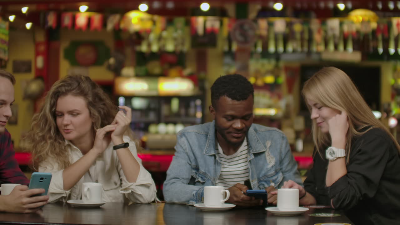 아름다운 히스패닉 남자는 멋진 스터 술집에서 웃는 농담과 술을 마시면서 친구들에게 스마트폰에서 흥미로운 것들을 보여줍니다.