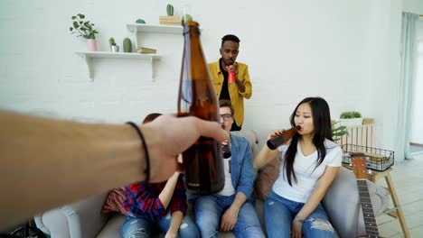 point of view shot of male hand clinking beer bottle with friends while celebrating party at home indoors