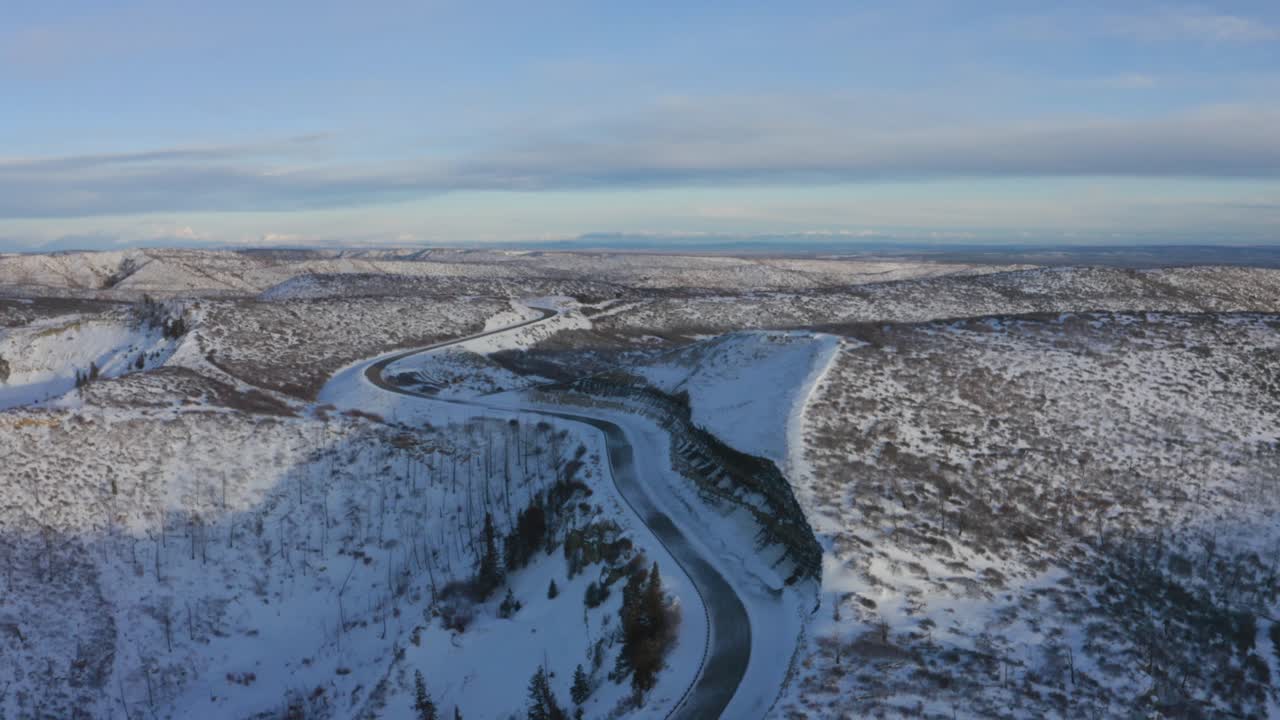 Vista aerea di una strada infinita che attraversa una catena montuosa innevata con una sommità pianeggiante coperta da vegetazione rada in una giornata di sole con nuvole in lontananza.