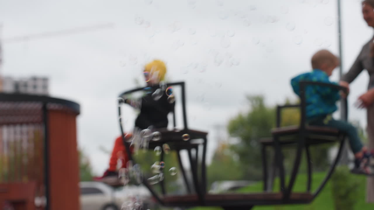 Blur background shows teacher spinning kindergarten children on merry go round while soap bubbles float across frame, soft focus railings and seats, cloudy sky over park