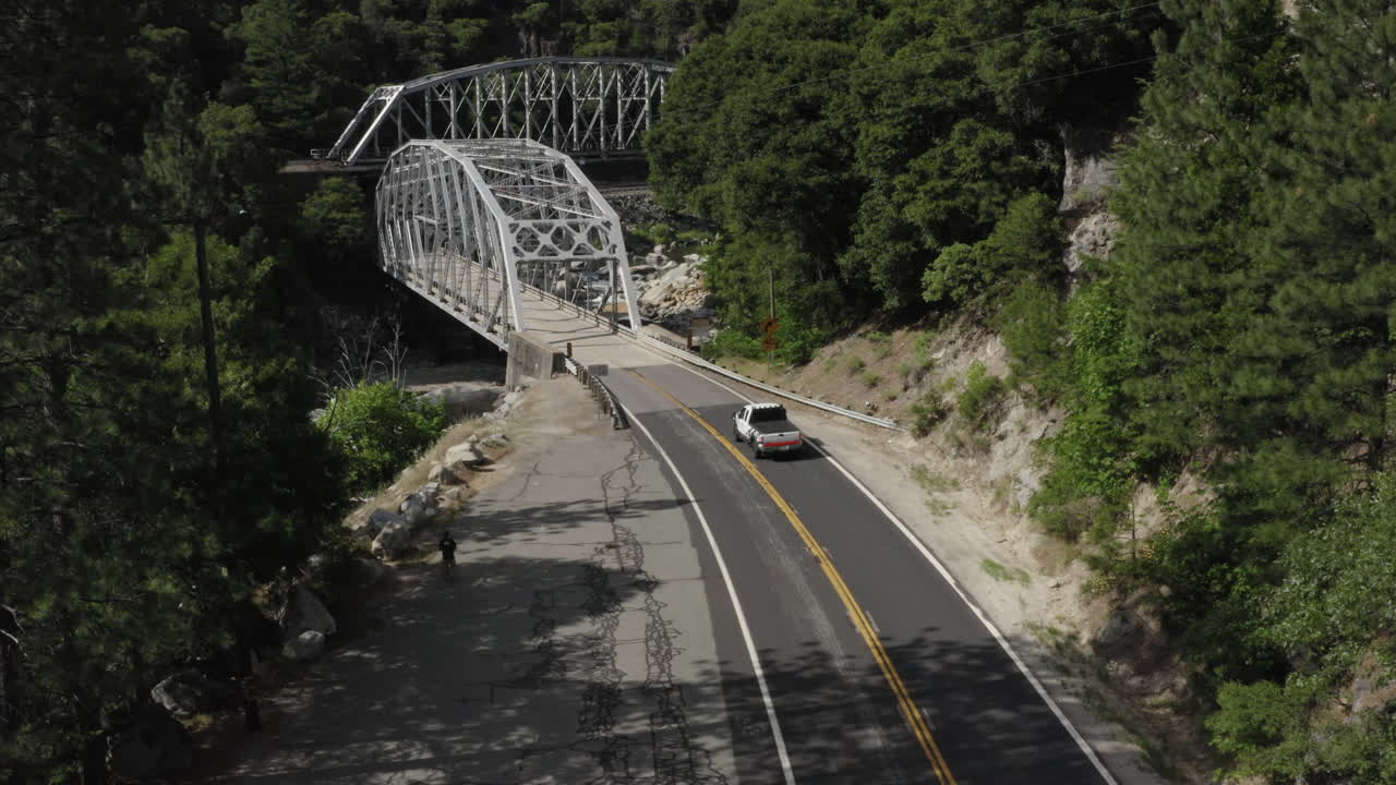 Lowering aerial above a road with white pick up truck passing below driving towards metal bridge on sunny day in national forest