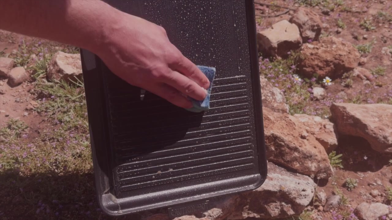 Arm reaches down to scrub ridged grill pan with sponge on rocky sunlit ground