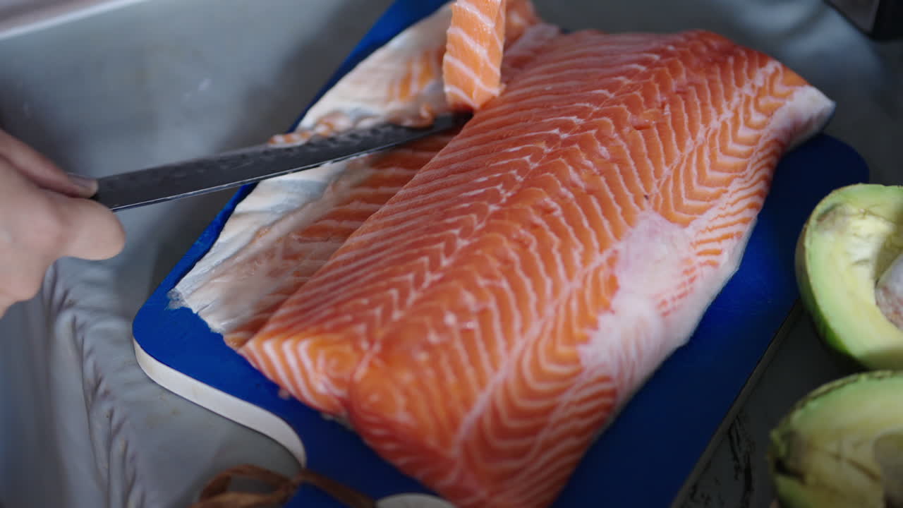 Top view of hands and knife slicing strip of fresh salmon fillet in kitchen
