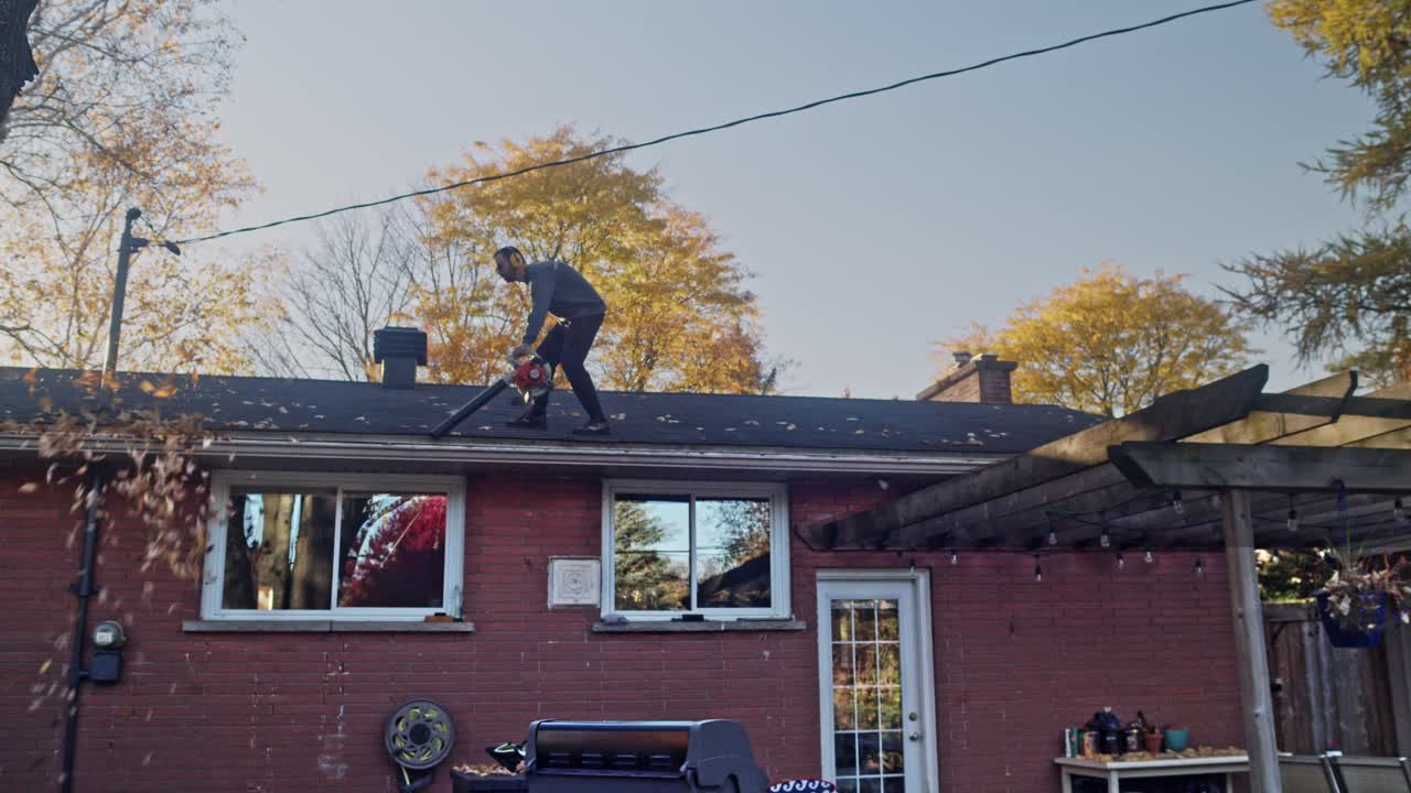 Man walks along a roof of a house with a gas leaf blower cleaning out of the gutters