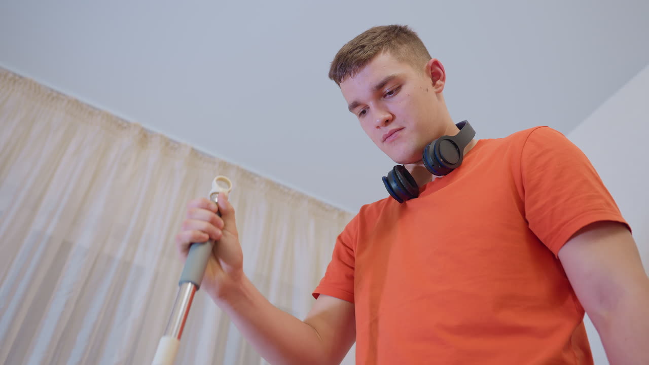Young boy in orange shirt with headphones around neck dips mop into water bucket and lifts it out preparing to mop floor during house cleaning