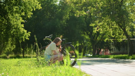 female cyclist crouches on grassy path under bright sunlight, pumping air into back tire of her bicycle, surrounded by lush greenery and trees, she works with focus