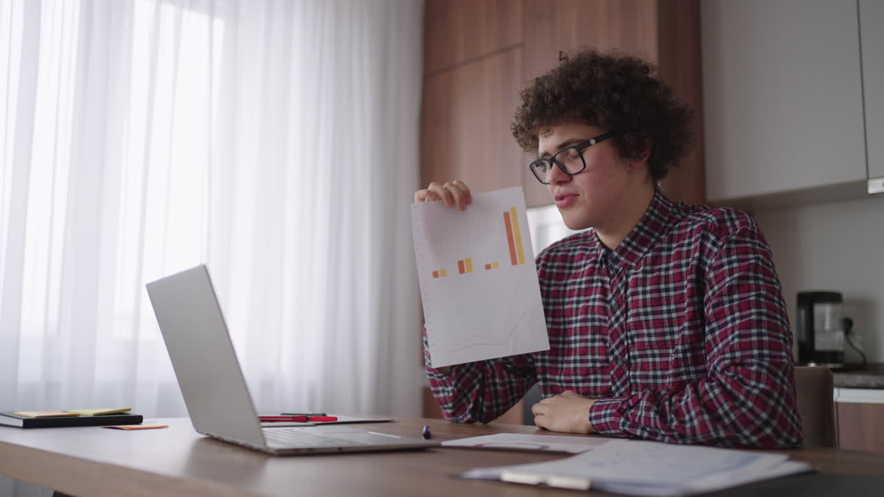 Curly - haired with glasses business man sitting at office from home desk looking at camera and pointing at a tablet with financial information displayed in graphical form column graph