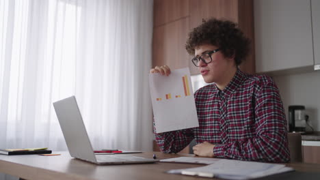 Curly---haired-with-glasses-business-man-sitting-at-office-from-home-desk-looking-at-camera-and-pointing-at-a-tablet-with-financial-information-displayed-in-graphical-form-column-graph