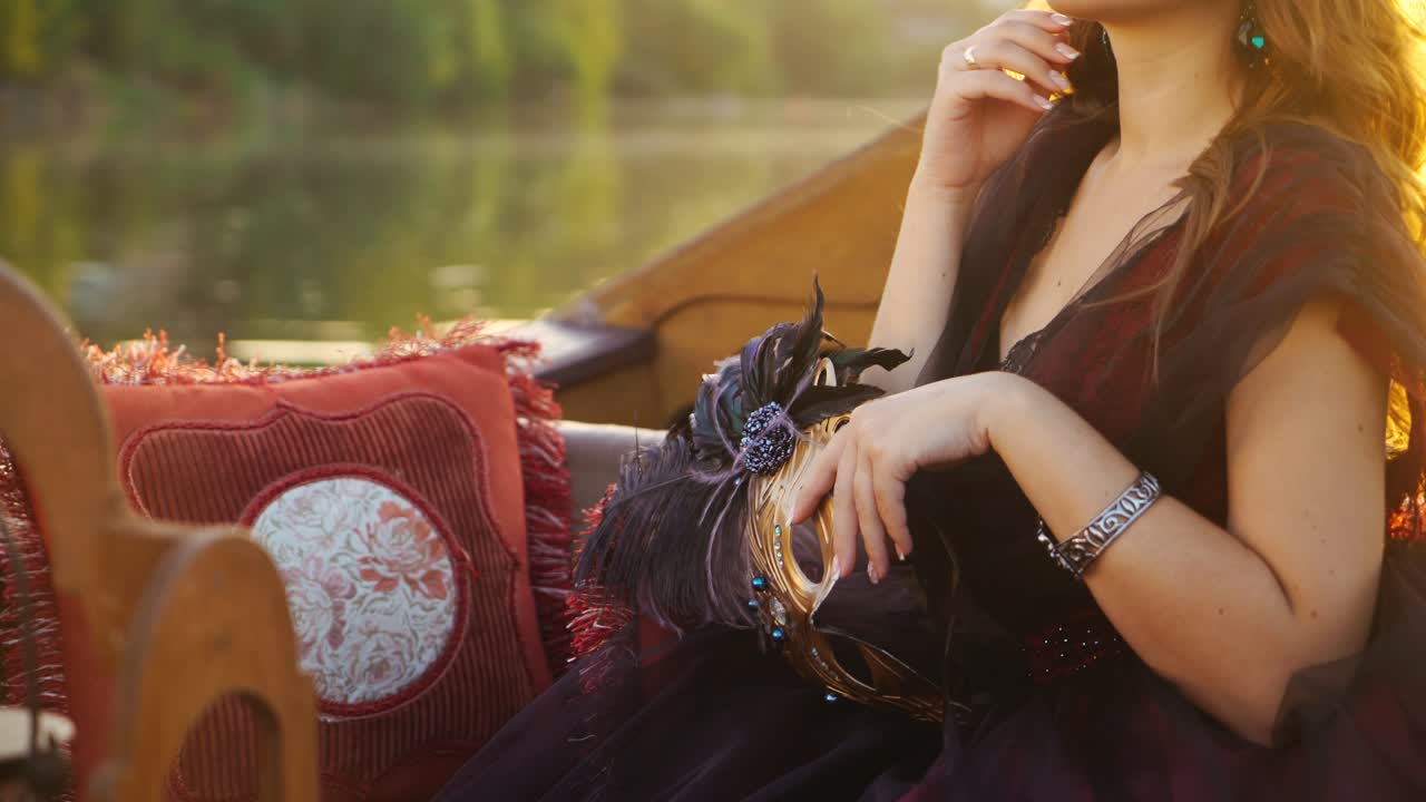 Luxury mask with feathers in the hand of a gorgeous woman in carnival dress floating in a boat at sunset. Attractive female in gondola with a mask in hand on the evening sun