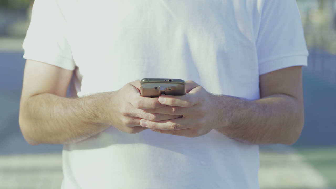 una foto recortada de un hombre con una camiseta blanca usando un teléfono inteligente.